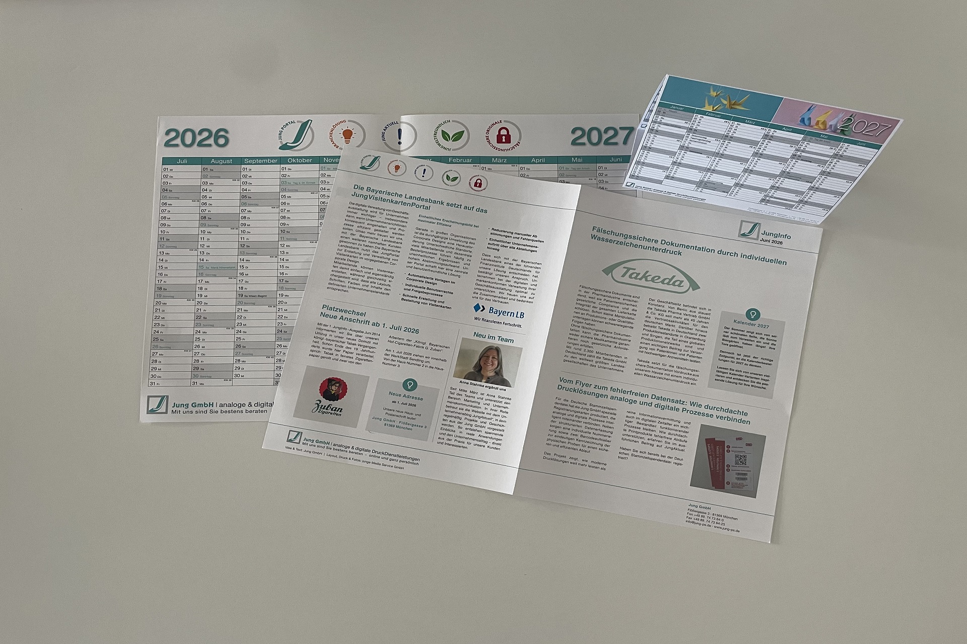 Auf einem hellen Hintergrund liegen verschiedene gedruckte Unternehmensmedien der Firma Jung GmbH. Zu sehen sind ein Wandkalender für die Jahre 2026 und 2027, ein gefalteter Newsletter mit mehreren Artikeln und Bildern sowie ein weiterer Kalenderflyer. Die Drucksachen präsentieren Unternehmensinformationen, Kundenprojekte, Mitarbeitervorstellungen und Termine und veranschaulichen die Leistungen des Unternehmens im Bereich analoge und digitale Druckdienstleistungen.
