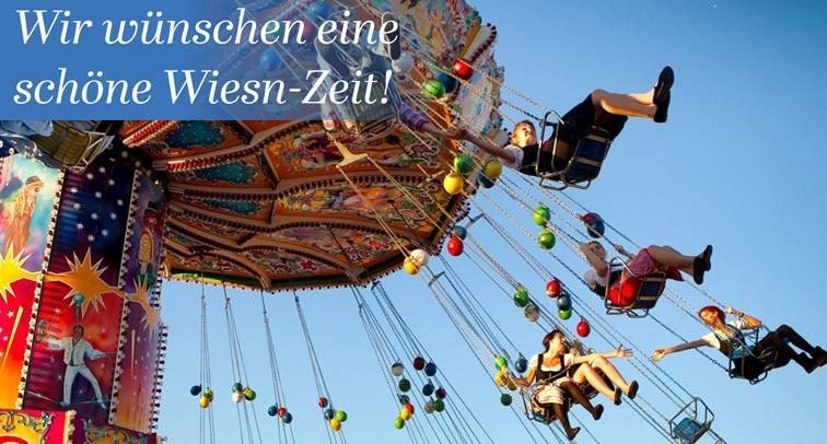 Buntes Kettenkarussell auf dem Münchner Oktoberfest mit fröhlichen Fahrgästen vor blauem Himmel. Im Bild steht: „Wir wünschen eine schöne Wiesn-Zeit“.